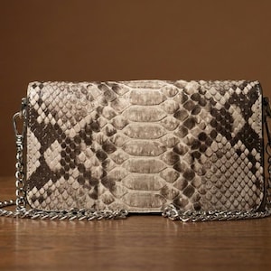 May include: A rectangular clutch bag featuring a textured snakeskin pattern in shades of beige, gray, and dark brown. The bag includes a detachable silver-toned metal chain strap attached to both sides with lobster claw clasps, providing a metallic finish.