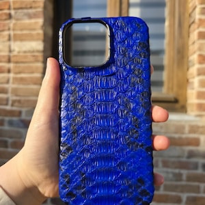 Puede incluir: Una funda de teléfono azul vibrante con un patrón texturizado en forma de escamas. La funda tiene un recorte para la cámara y se sostiene contra un fondo de ladrillo. La funda parece estar hecha de un material que imita la piel de serpiente.
