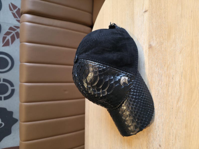 May include: Black baseball cap with a snakeskin-patterned visor and accents. The cap features a suede crown and a small silver emblem on the side. The hat is displayed on a wooden surface.