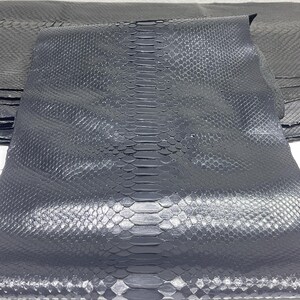 Op de afbeelding: Close-up van donkergrijs slangenleer. Het leer heeft een getextureerd, geschubd patroon met een prominente centrale ruggengraat. Het materiaal lijkt gevouwen, waardoor de onderkant en de randen van het leer zichtbaar worden. De belichting benadrukt de textuur en kleurvariaties.