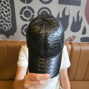 以下が含まれることがあります： 黒い野球帽、ヘビ革のような質感。 つばがカーブしており、クラウンは構造化されています。 帽子は暗い素材でできており、スタイリッシュなアクセサリーです。