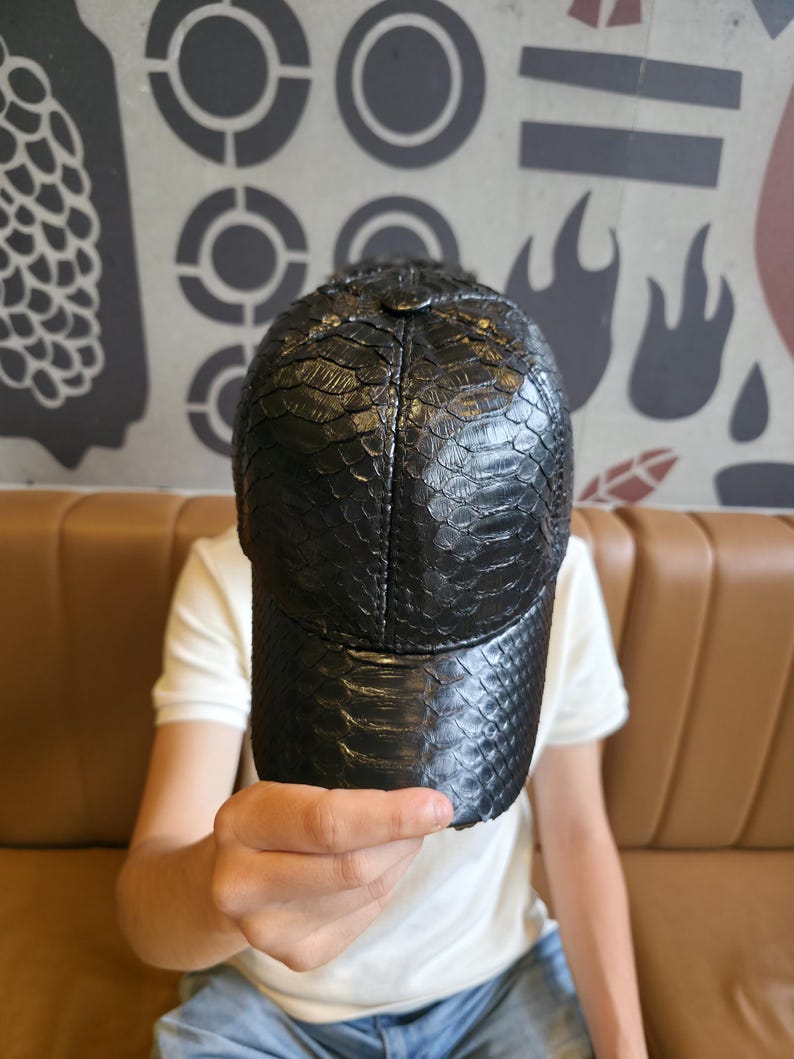 K&ouml;nnte beinhalten: Schwarze Baseballkappe mit einem strukturierten, schlangenlederartigen Muster. Die Kappe hat einen gebogenen Schirm und eine strukturierte Krone. Der Hut ist aus dunklem Material gefertigt und ein stilvolles Accessoire.