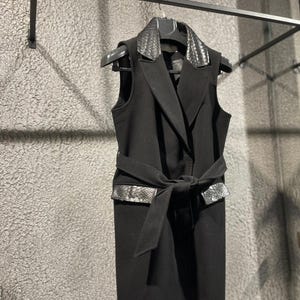 Könnte beinhalten: Eine schwarze ärmellose Weste mit Kunstschlangenkragen und Gürteldetails. Die Weste hängt an einem schwarzen Kleiderbügel. Der Gürtel ist vorne gebunden.