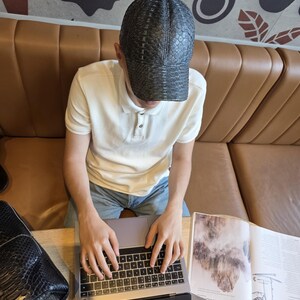 May include: A black cap with a snakeskin pattern is worn by a person sitting at a table. The person is wearing a white polo shirt and blue jeans. A laptop and a black bag are on the table.
