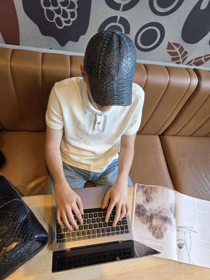 May include: A black cap with a snakeskin pattern is worn by a person sitting at a table. The person is wearing a white polo shirt and blue jeans. A laptop and a black bag are on the table.