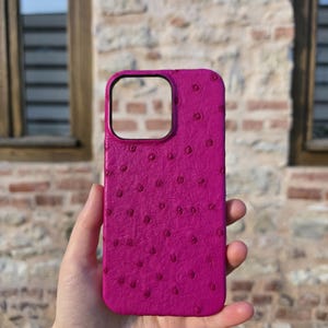 Pode incluir: Uma capa de telefone magenta com um padrão texturizado e pontilhado. A capa tem uma forma retangular com um recorte para a câmera. O fundo é uma parede de tijolos e uma janela.