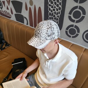 May include: A white and gray snakeskin-patterned baseball cap. The cap is worn by a person sitting on a brown leather couch and reading a book. The person is wearing a white polo shirt and blue jeans.