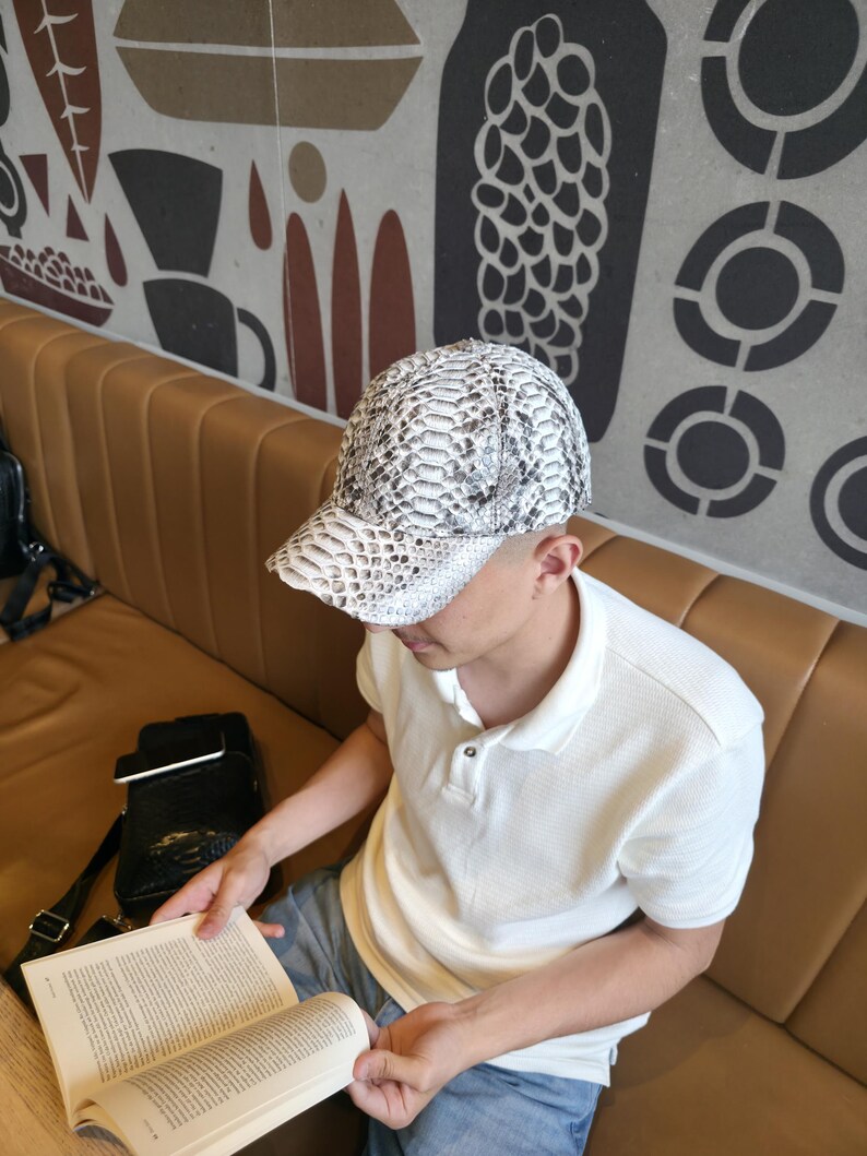 May include: A white and gray snakeskin-patterned baseball cap. The cap is worn by a person sitting on a brown leather couch and reading a book. The person is wearing a white polo shirt and blue jeans.