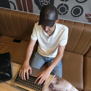 Op de afbeelding: Een bruine, getextureerde baseballcap met een gebogen klep. De pet wordt gedragen door een persoon die een witte poloshirt en een blauwe spijkerbroek draagt. Een laptop en een tijdschrift liggen op een houten tafel.