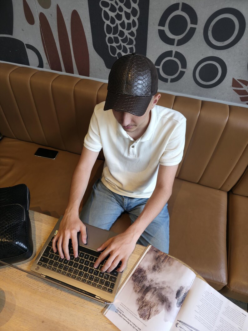 Op de afbeelding: Een bruine, getextureerde baseballcap met een gebogen klep. De pet wordt gedragen door een persoon die een witte poloshirt en een blauwe spijkerbroek draagt. Een laptop en een tijdschrift liggen op een houten tafel.