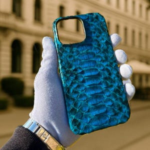 Op de afbeelding: Een felblauwe telefoonhoes met een slangenleertextuur. De hoes heeft een rechthoekige vorm met afgeronde hoeken en uitsparingen voor de camera en knoppen. De hoes wordt vastgehouden door een gehandschoende hand, die het patroon en de kleur laat zien.