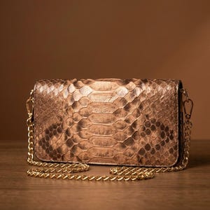 May include: A rectangular metallic bronze clutch bag featuring a textured snakeskin pattern. The bag includes a detachable gold-tone metal chain shoulder strap attached to side clasps, providing a structured appearance with a shimmering finish.