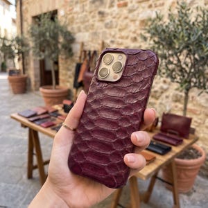 May include: A deep burgundy smartphone case featuring a textured, scale-like pattern across the entire back surface. The case includes a precise cutout for a triple-lens camera array, showcasing the metallic rings around each individual lens.