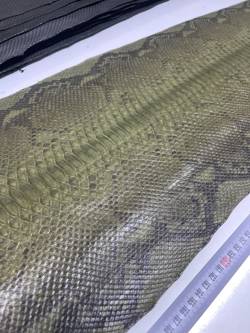 Peut inclure: Gros plan sur un motif peau de serpent dans des tons verts et gris. La surface textur&eacute;e pr&eacute;sente un motif en forme d'&eacute;cailles. Un mat&eacute;riau noir est visible dans le coin sup&eacute;rieur gauche. Une r&egrave;gle avec des marquages en centim&egrave;tres est visible.