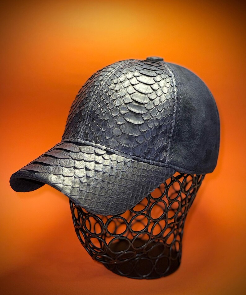 May include: A dark blue baseball cap with a textured, scaly pattern on the front panels and bill. The back of the cap is a smooth, dark blue suede material. The cap is displayed on a black wire form against an orange background.