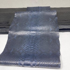 Könnte beinhalten: Nahaufnahme von blauen und schwarzen Pythonlederplatten. Das Leder hat ein Schuppenmuster und eine glänzende Oberfläche. Die Platten sind übereinander gestapelt, wobei die oberste Platte leicht gefaltet ist. Der Hintergrund ist eine schlichte weiße Oberfläche.