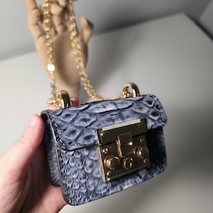 May include: A miniature handbag with a blue and grey snakeskin pattern and a gold chain strap. It features a gold clasp. The bag is held in a hand.