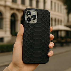 May include: Black phone case featuring a snakeskin texture. The case has a square camera module with three lenses and a flash. The phone case is held in a hand, with a blurred background of a building and street.