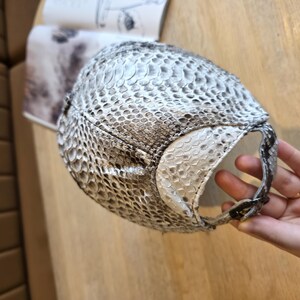 May include: A baseball cap with a snakeskin pattern in shades of white, gray, and black. The cap has a curved brim and an adjustable strap. The cap is made of a textured material.