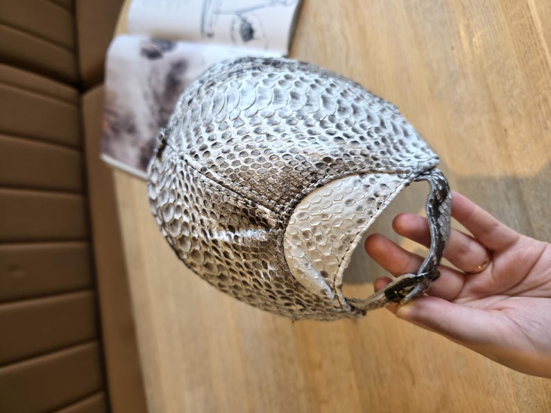 May include: A baseball cap with a snakeskin pattern in shades of white, gray, and black. The cap has a curved brim and an adjustable strap. The cap is made of a textured material.