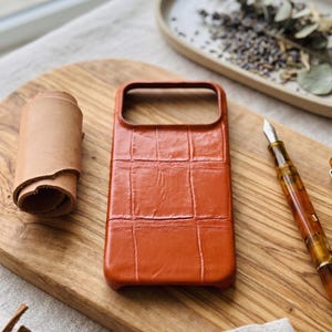 May include: An orange leather phone case with a crocodile skin texture rests on a wooden board. A roll of tan leather and a fountain pen are also present. A tray with dried plants is in the background.