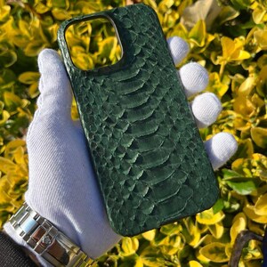 Könnte beinhalten: Dunkelgrüne Handyhülle mit Schlangenhautstruktur. Die Hülle wird von einer behandschuhten Hand gehalten, die das detaillierte Muster und die Farbe hervorhebt. Die Hülle ist für ein Smartphone konzipiert.