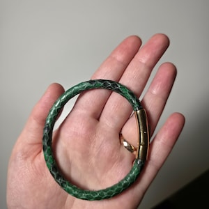 Peut inclure: Un bracelet en peau de serpent vert et noir avec un fermoir doré. Le bracelet est circulaire et semble être en cuir véritable. Le bracelet est tenu dans la main d'une personne.