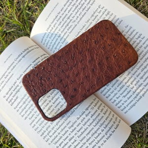 Pode incluir: Uma capa de telefone de couro marrom com uma superf&iacute;cie texturizada repousa sobre um livro aberto. A capa tem um recorte para a c&acirc;mera e uma apar&ecirc;ncia lisa e polida. As p&aacute;ginas do livro est&atilde;o cheias de texto.