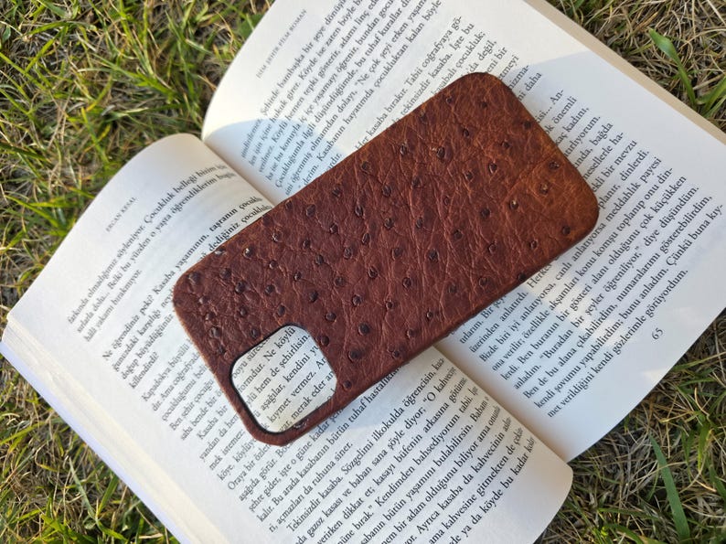Pode incluir: Uma capa de telefone de couro marrom com uma superf&iacute;cie texturizada repousa sobre um livro aberto. A capa tem um recorte para a c&acirc;mera e uma apar&ecirc;ncia lisa e polida. As p&aacute;ginas do livro est&atilde;o cheias de texto.