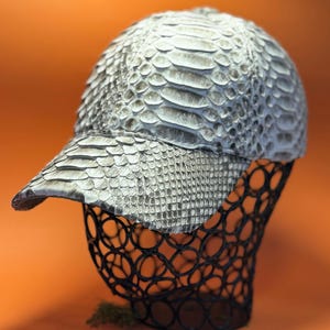 Può includere: Un cappellino da baseball con un motivo strutturato e squamoso nei toni del grigio e del bianco. Il cappellino è esposto su una forma di filo nero su sfondo arancione. La visiera si estende in avanti e la superficie del cappellino ha una consistenza simile a quella di un rettile.