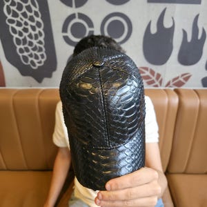 K&ouml;nnte beinhalten: Schwarze Baseballkappe mit einem strukturierten, schlangenlederartigen Muster. Die Kappe hat einen gebogenen Schirm und einen Knopf oben. Der Hut wird vor einer Person gehalten.