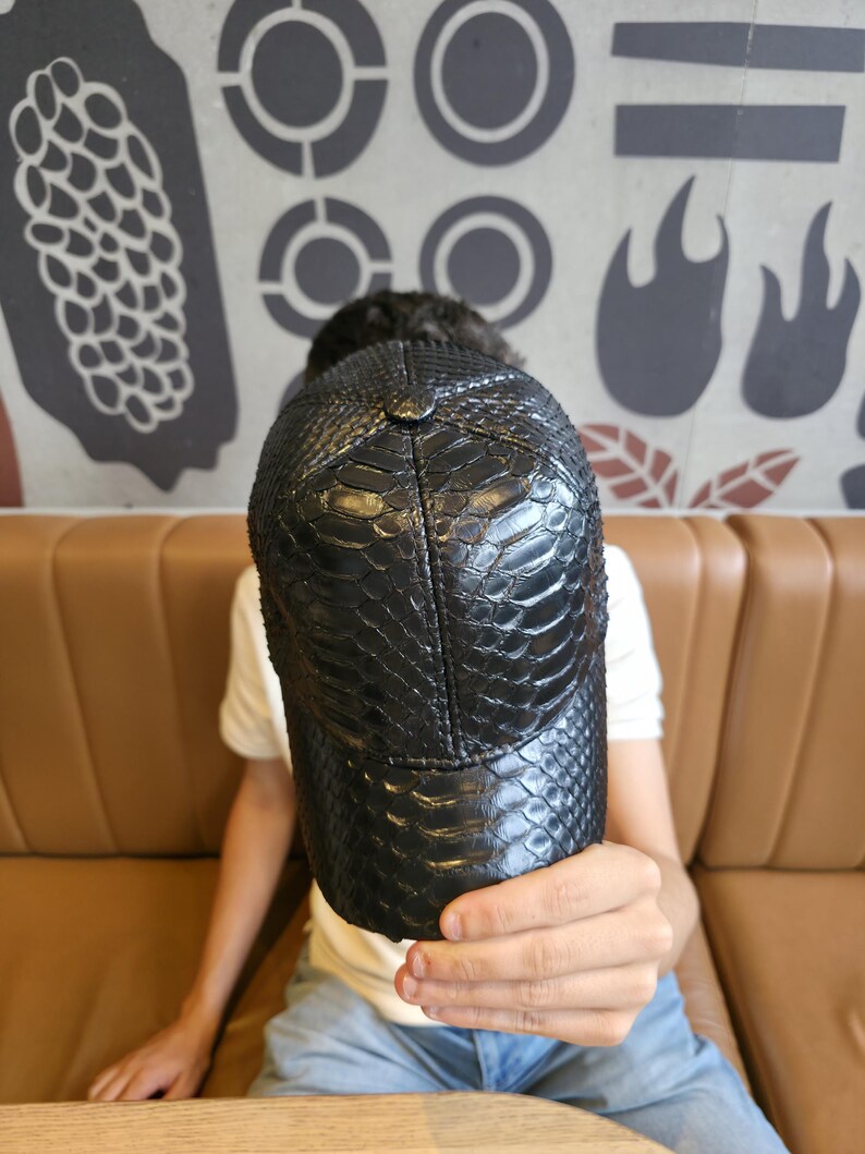 K&ouml;nnte beinhalten: Schwarze Baseballkappe mit einem strukturierten, schlangenlederartigen Muster. Die Kappe hat einen gebogenen Schirm und einen Knopf oben. Der Hut wird vor einer Person gehalten.