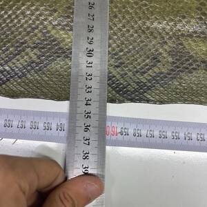 Peut inclure: Gros plan sur un mat&eacute;riau textur&eacute; vert olive et noir. Une r&egrave;gle m&eacute;tallique est plac&eacute;e verticalement, affichant des mesures en centim&egrave;tres. La r&egrave;gle est positionn&eacute;e sur le mat&eacute;riau, avec les chiffres 23 &agrave; 39 visibles.