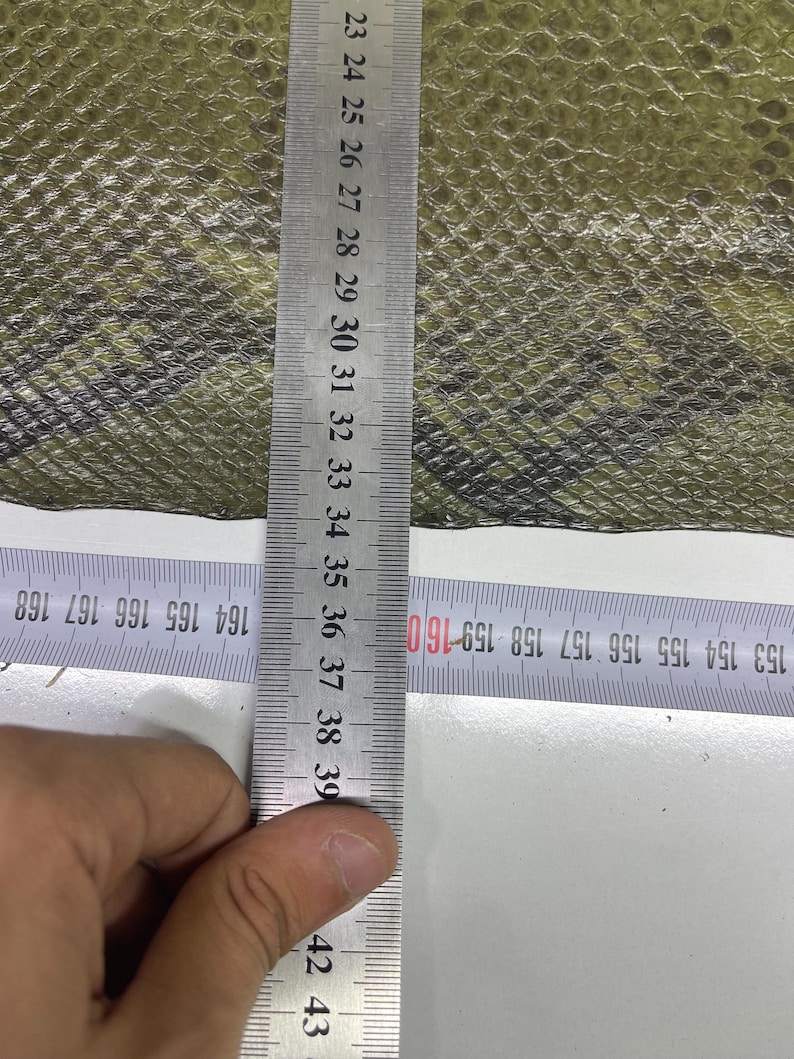 Peut inclure: Gros plan sur un mat&eacute;riau textur&eacute; vert olive et noir. Une r&egrave;gle m&eacute;tallique est plac&eacute;e verticalement, affichant des mesures en centim&egrave;tres. La r&egrave;gle est positionn&eacute;e sur le mat&eacute;riau, avec les chiffres 23 &agrave; 39 visibles.