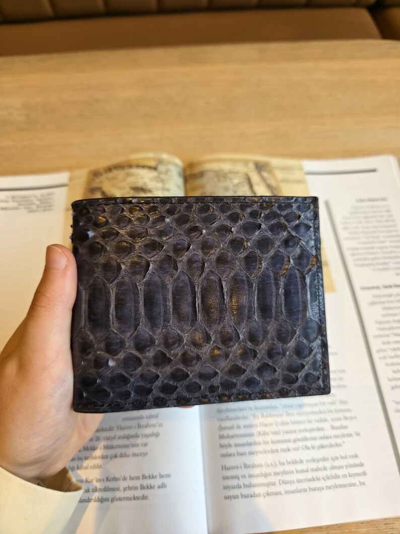 Puede incluir: Una cartera azul oscuro con un patr&oacute;n texturizado en forma de escamas. La cartera rectangular parece estar hecha de piel de reptil. La cartera se sostiene abierta, revelando el interior.