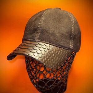 May include: A brown baseball cap with a textured, patterned brim. The cap is displayed on a black wire form against an orange background. The cap's crown is a darker brown, with visible stitching.