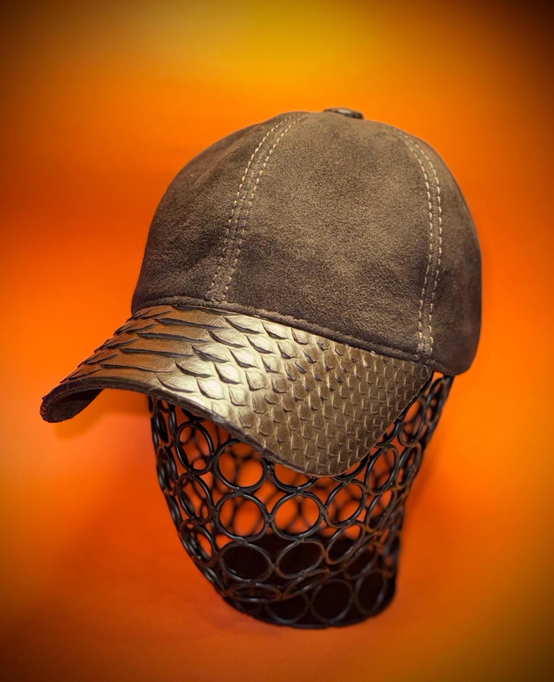 May include: A brown baseball cap with a textured, patterned brim. The cap is displayed on a black wire form against an orange background. The cap's crown is a darker brown, with visible stitching.