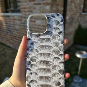 Op de afbeelding: Een telefoonhoesje met een wit en grijs slangenpatroon. De hoes heeft een rechthoekige vorm met een uitsparing voor de camera en een glad, gestructureerd oppervlak. Het ontwerp bootst het uiterlijk van slangenhuid na.