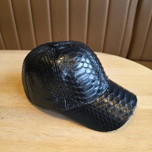 K&ouml;nnte beinhalten: Schwarze Baseballkappe mit einem strukturierten, reptilienartigen Muster. Die Kappe hat einen gebogenen Schirm und eine abgerundete Krone. Der Hut besteht aus einem Material, das Schlangenhaut &auml;hnelt. Die Kappe liegt auf einer hellbraunen Holzoberfl&auml;che.