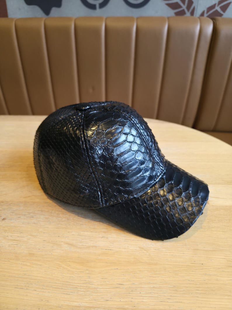 K&ouml;nnte beinhalten: Schwarze Baseballkappe mit einem strukturierten, reptilienartigen Muster. Die Kappe hat einen gebogenen Schirm und eine abgerundete Krone. Der Hut besteht aus einem Material, das Schlangenhaut &auml;hnelt. Die Kappe liegt auf einer hellbraunen Holzoberfl&auml;che.