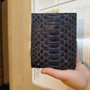 Puede incluir: Una cartera azul oscuro con un patr&oacute;n de piel de serpiente. La cartera es rectangular y parece estar hecha de cuero. La textura de la piel de serpiente es visible, con un dise&ntilde;o similar a escamas. La cartera se sostiene en una mano.