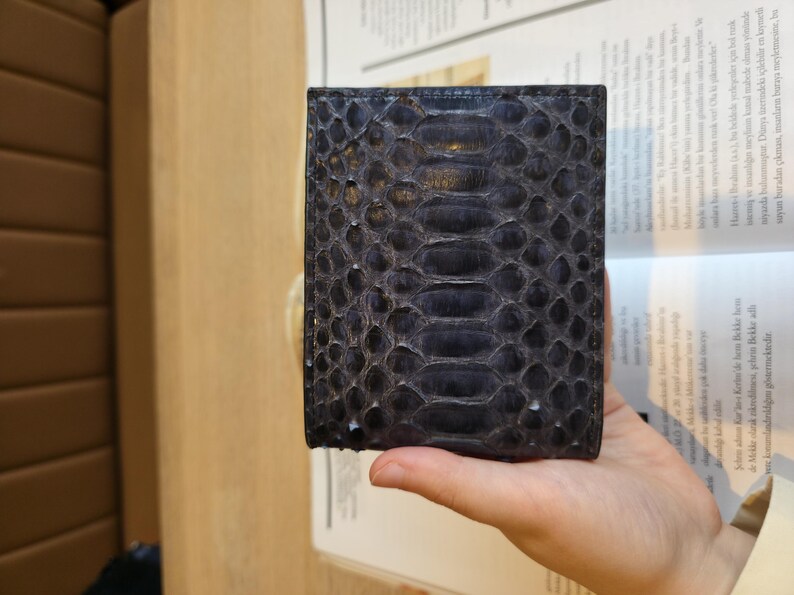 Puede incluir: Una cartera azul oscuro con un patr&oacute;n de piel de serpiente. La cartera es rectangular y parece estar hecha de cuero. La textura de la piel de serpiente es visible, con un dise&ntilde;o similar a escamas. La cartera se sostiene en una mano.