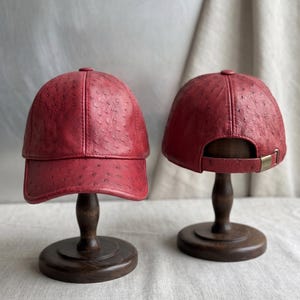 May include: Two red baseball caps with a textured ostrich leather appearance are displayed on wooden hat stands. The caps have a curved brim and a brass-coloured adjustable strap. The stands are dark brown with a round base.