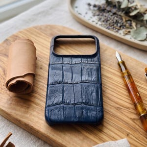 Puede incluir: Una funda de teléfono azul marino con textura de piel de cocodrilo está sobre una tabla de cortar de madera. Un rollo de cuero marrón claro y una pluma estilográfica también están en la tabla. La funda está diseñada para proteger un smartphone.