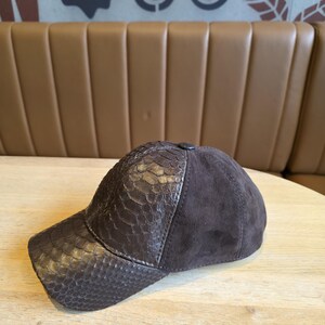 Op de afbeelding: Een donkerbruine baseballcap met een klep met slangenleertextuur en een su&egrave;de kroon. De pet heeft een knoop aan de bovenkant. De hoed ligt op een lichtgekleurd houten oppervlak.
