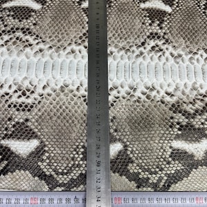 以下が含まれることがあります： グレー、ブラウン、白の鱗模様のヘビ革。金属製の定規が革の上に垂直に配置され、センチメートル単位の測定値が表示されています。革の中央には白いストライプがあります。