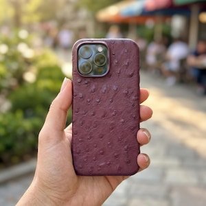 Puede incluir: Una funda para smartphone de color burdeos oscuro con un patrón de cuero de avestruz texturizado y sus características protuberancias. La funda tiene un recorte rectangular en la esquina superior izquierda para una cámara triple.