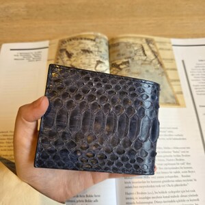 Puede incluir: Una cartera de cuero azul con un patr&oacute;n de piel de serpiente. La cartera es rectangular y parece estar doblada. La cartera est&aacute; sujeta en la mano de una persona. El fondo incluye un libro y una superficie de madera.