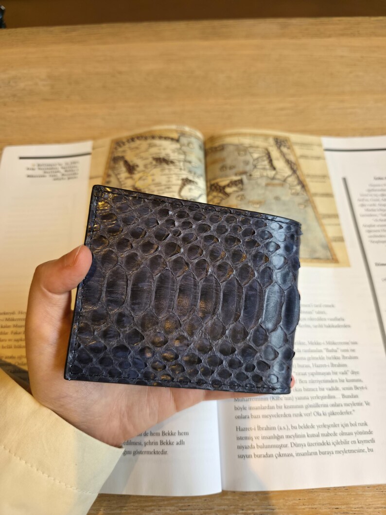 Puede incluir: Una cartera de cuero azul con un patr&oacute;n de piel de serpiente. La cartera es rectangular y parece estar doblada. La cartera est&aacute; sujeta en la mano de una persona. El fondo incluye un libro y una superficie de madera.