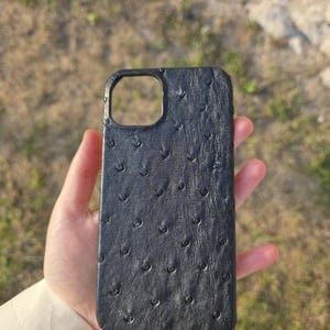 Könnte beinhalten: Schwarze Handyhülle mit strukturierter, genoppter Oberfläche. Die Hülle hat einen Ausschnitt für die Kamera und ist für ein Smartphone konzipiert. Die Hülle besteht aus einem dunklen Material, möglicherweise Leder oder einem lederähnlichen Material.
